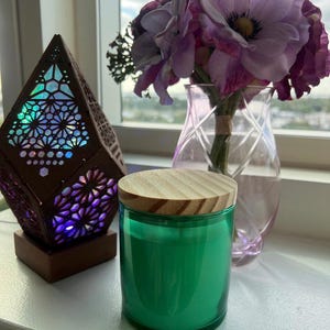 May include: A green glass candle with a wooden lid sits on a white surface next to a decorative wooden lamp with a colorful stained glass design. A vase with purple flowers is in the background.