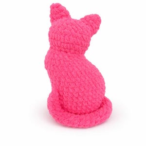 May include: A bright pink, handmade cat-shaped plush toy. The cat is sitting upright with its tail curled and ears perked up. The plush has a textured, fuzzy appearance and is set against a white background.