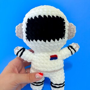 May include: White crocheted astronaut toy with black eyes and a red and blue patch on the chest. The toy is held in a hand with pink and black nail polish.