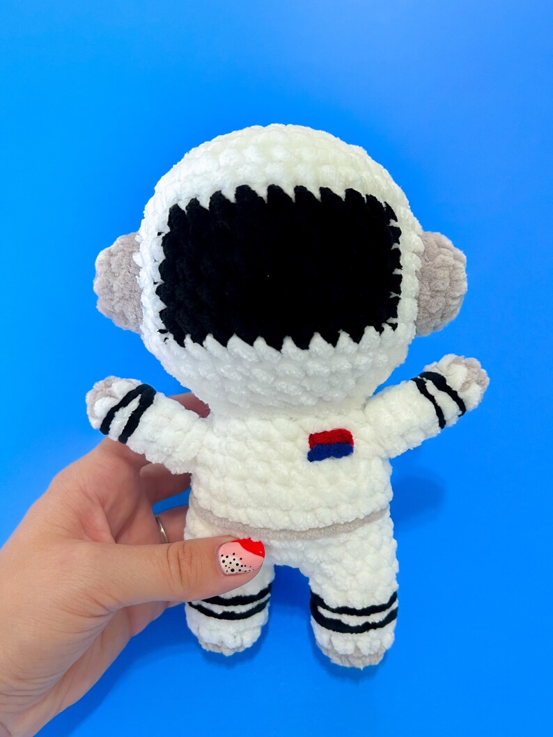 May include: White crocheted astronaut toy with black eyes and a red and blue patch on the chest. The toy is held in a hand with pink and black nail polish.