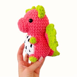 Puede incluir: Un juguete de drag&oacute;n de ganchillo hecho a mano en rosa y verde. El drag&oacute;n tiene ojos negros, alas verdes y detalles verdes en la cabeza y los pies. El vientre es blanco con manchas negras, que se asemejan a una fruta del drag&oacute;n.
