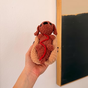 Op de afbeelding: Een handgemaakt gehaakt hondenspeeltje in een hotdogbroodje. De hond is bruin met een rode lijn en het broodje is bruin. Het speelgoed wordt in een hand gehouden, met een witte muur en een abstract schilderij met lijst op de achtergrond.
