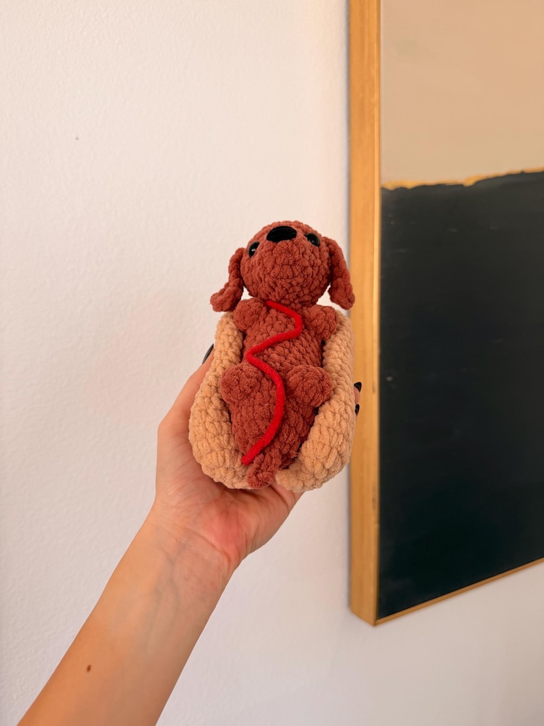 Op de afbeelding: Een handgemaakt gehaakt hondenspeeltje in een hotdogbroodje. De hond is bruin met een rode lijn en het broodje is bruin. Het speelgoed wordt in een hand gehouden, met een witte muur en een abstract schilderij met lijst op de achtergrond.