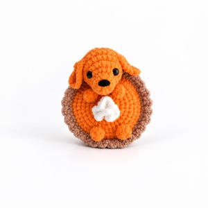May include: A crocheted stuffed animal shaped like a small orange dog sitting inside a round orange pie with a brown crust and a white dollop of whipped cream on top. The dog has black bead eyes and a black nose, with floppy ears and small paws.
