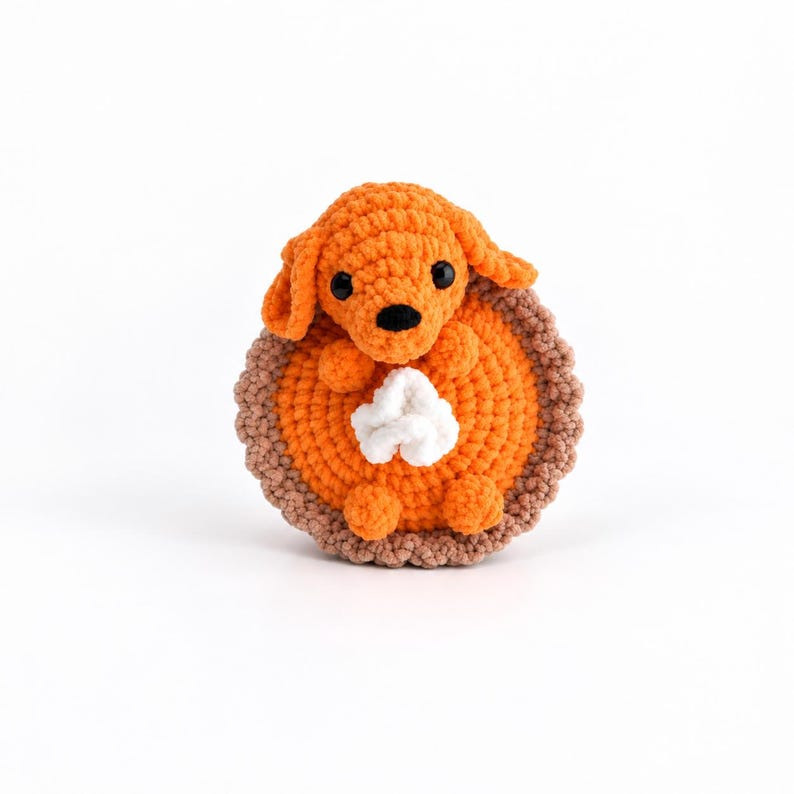 May include: A crocheted stuffed animal shaped like a small orange dog sitting inside a round orange pie with a brown crust and a white dollop of whipped cream on top. The dog has black bead eyes and a black nose, with floppy ears and small paws.
