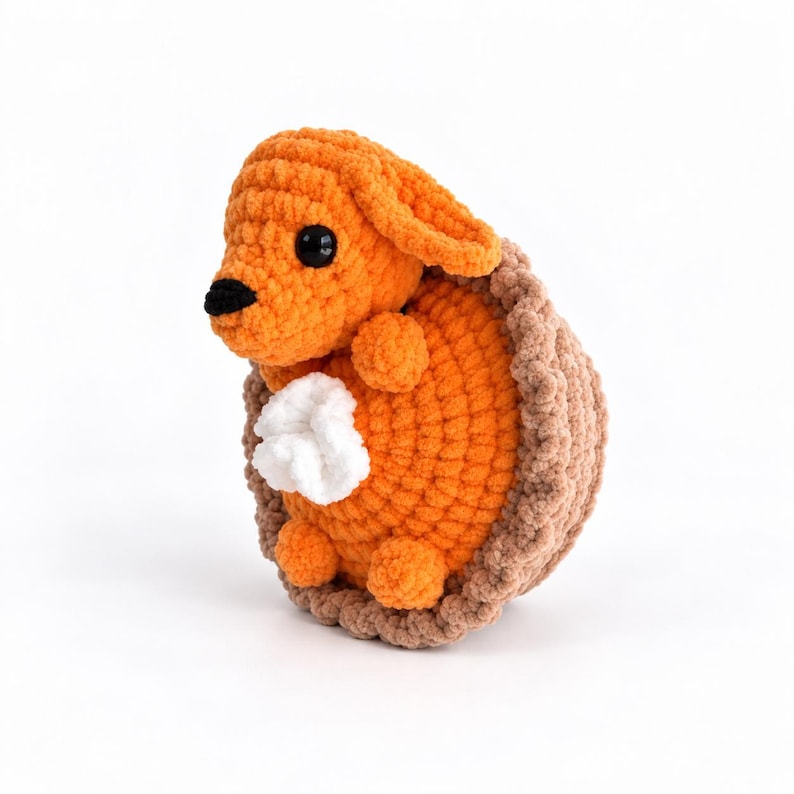 May include: A crocheted stuffed animal featuring an orange creature with black eyes and a black nose, nestled inside a brown textured shell-like structure. A small white tuft of yarn is attached to the center of the orange body.