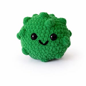 May include: A handmade, green crocheted plushie in the shape of a round, spiky ball. The plushie has two large, black, button eyes and a stitched smile. The toy is made of a soft, textured yarn.