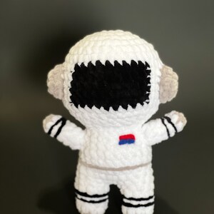 May include: White crocheted astronaut toy with black eyes, a red and blue patch on the chest, and black and white stripes on the legs.