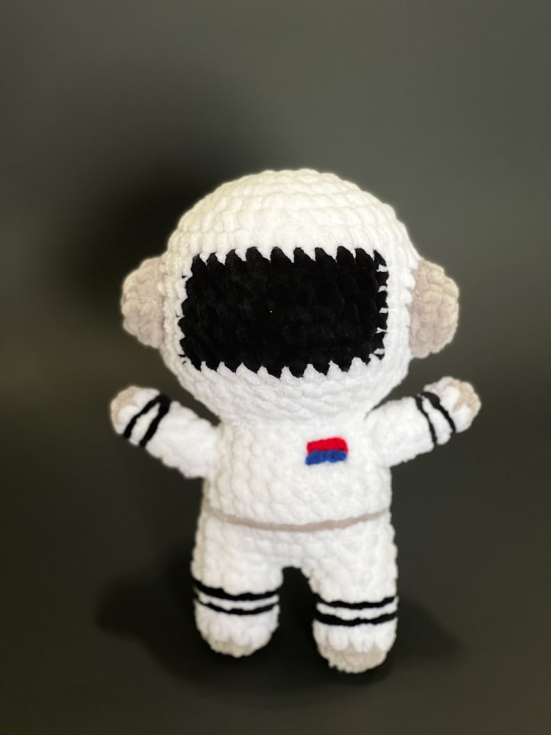 May include: White crocheted astronaut toy with black eyes, a red and blue patch on the chest, and black and white stripes on the legs.