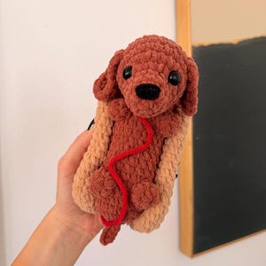Op de afbeelding: Een handgemaakte gehaakte hond in een hotdogbroodje. De hond is bruin met zwarte ogen en een rode lijn. Het hotdogbroodje is bruin en beige. Het speelgoed is zacht en pluche.