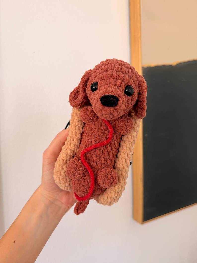 Op de afbeelding: Een handgemaakte gehaakte hond in een hotdogbroodje. De hond is bruin met zwarte ogen en een rode lijn. Het hotdogbroodje is bruin en beige. Het speelgoed is zacht en pluche.