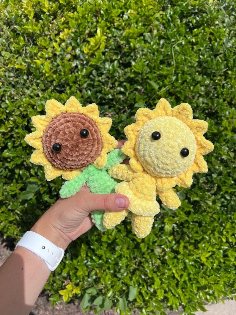 May include: Two crocheted sunflower plushies, one brown and one yellow, with black eyes. The plushies are held in a hand against a green leafy background.