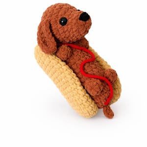 Op de afbeelding: Een bruine gehaakte teckel met zwarte ogen en een rode lijn, genesteld in een gehaakt, beige hotdogbroodje. Het pluche speelgoed staat tegen een witte achtergrond en cre&euml;ert een speelse en grillige esthetiek.