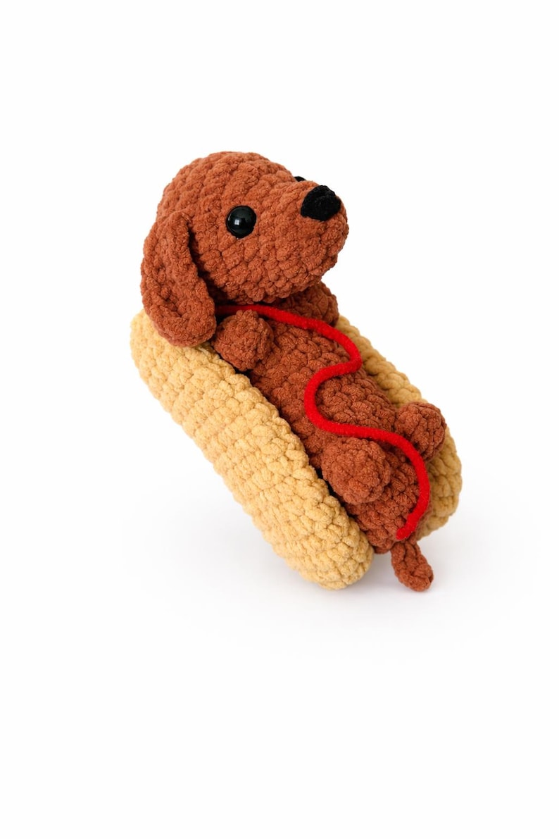 Op de afbeelding: Een bruine gehaakte teckel met zwarte ogen en een rode lijn, genesteld in een gehaakt, beige hotdogbroodje. Het pluche speelgoed staat tegen een witte achtergrond en cre&euml;ert een speelse en grillige esthetiek.