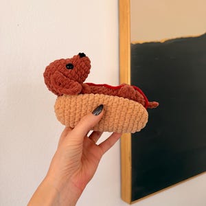Op de afbeelding: Een handgemaakt gehaakt hotdog-speeltje. De hotdog is bruin, met een rode 'ketchup'-accent, genesteld in een beige broodje. Het speeltje wordt in een hand gehouden. De achtergrond omvat een witte muur en een ingelijst kunstwerk.