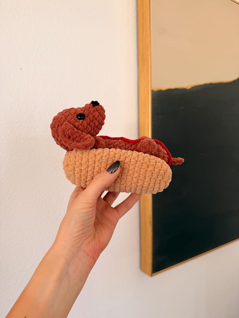 Op de afbeelding: Een handgemaakt gehaakt hotdog-speeltje. De hotdog is bruin, met een rode 'ketchup'-accent, genesteld in een beige broodje. Het speeltje wordt in een hand gehouden. De achtergrond omvat een witte muur en een ingelijst kunstwerk.