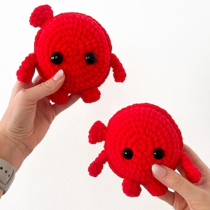 May include: Two red crocheted plush toys shaped like a crab with black eyes. The toys are being held by hands.