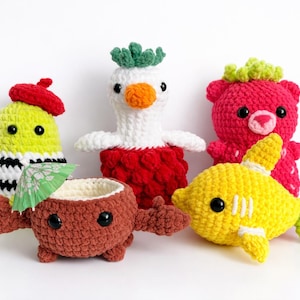 May include: Five brightly coloured crochet toys are displayed. They include a yellow creature with a red beret, a white and red bird-like figure, a red bear, a brown coconut with a tiny umbrella, and a yellow fish.