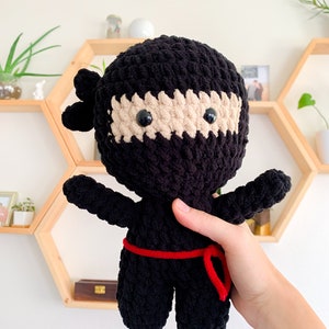 May include: Black crocheted ninja plush toy with a beige face and red sash. The toy is being held by a hand.