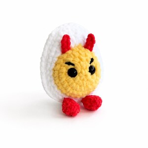 May include: A crocheted plush toy shaped like a hard-boiled egg with a white outer layer and a yellow center. It features two small red horns on top, two black plastic bead eyes, stitched black eyebrows, and two small red feet at the base.