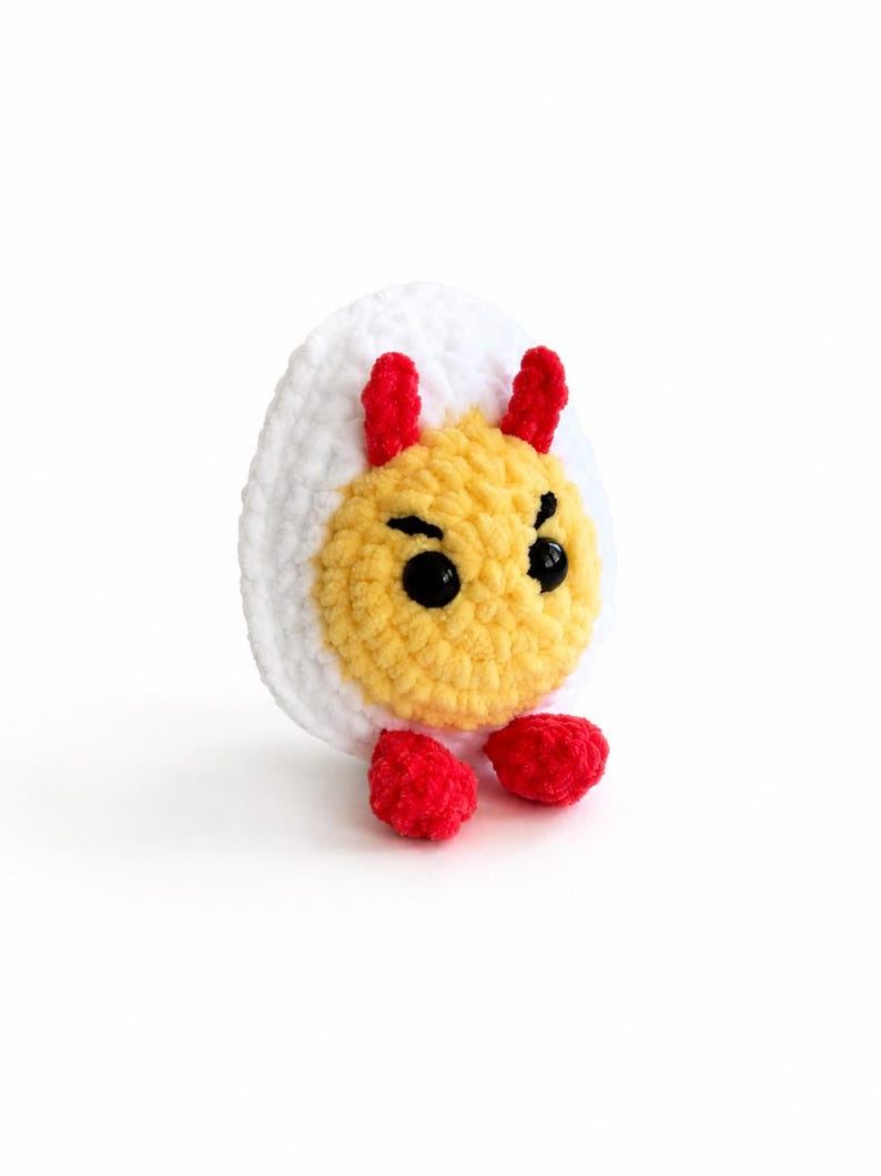 May include: A crocheted plush toy shaped like a hard-boiled egg with a white outer layer and a yellow center. It features two small red horns on top, two black plastic bead eyes, stitched black eyebrows, and two small red feet at the base.