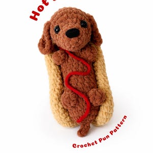 Op de afbeelding: Een handgemaakte gehaakte hotdog met een bruine hond in een bruin broodje, met een rood touwtje. De woorden "Hot Dog" en "Crochet Pun Pattern" zijn in het rood geschreven.