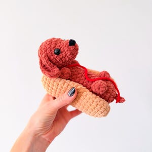 Op de afbeelding: Een handgemaakt gehaakt speelgoed van een teckel in een hotdogbroodje. De hond is roodbruin met zwarte ogen en neus, en een rode touwketting. Het broodje is bruin.