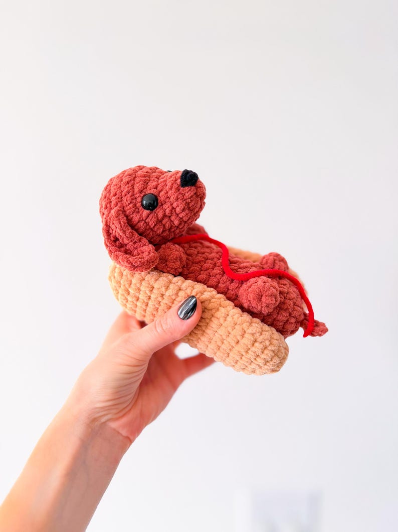 Op de afbeelding: Een handgemaakt gehaakt speelgoed van een teckel in een hotdogbroodje. De hond is roodbruin met zwarte ogen en neus, en een rode touwketting. Het broodje is bruin.