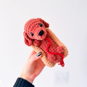 Op de afbeelding: Een handgemaakte gehaakte hond in een hotdogbroodje. De hond is roodbruin met zwarte ogen en een zwarte neus. Een rood garen loopt van de hond. Het broodje is bruin. De hond is een grillig, speels ontwerp.