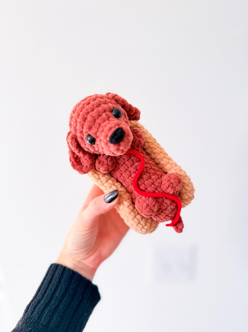 Op de afbeelding: Een handgemaakte gehaakte hond in een hotdogbroodje. De hond is roodbruin met zwarte ogen en een zwarte neus. Een rood garen loopt van de hond. Het broodje is bruin. De hond is een grillig, speels ontwerp.