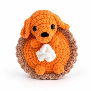 May include: A crocheted orange dog plushie sitting inside a round orange pie with a brown crust edge and a white dollop of whipped cream on top. The dog has black plastic eyes and a black nose, with floppy ears and small paws resting on the pie.