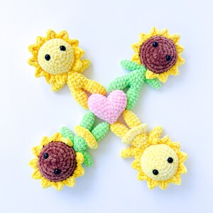 May include: Four crocheted sunflower plushies holding a pink heart in the center. The plushies are made with yellow, brown, and green yarn.