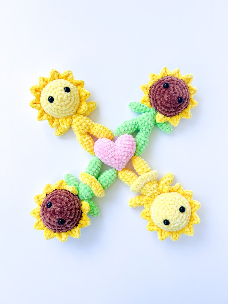May include: Four crocheted sunflower plushies holding a pink heart in the center. The plushies are made with yellow, brown, and green yarn.