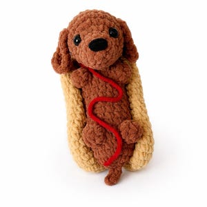 Puede incluir: Un juguete de perro de ganchillo marrón hecho a mano con ojos y nariz negros, metido en un pan de hot dog de ganchillo beige. Una correa roja de ganchillo se extiende desde el perro. Sobre un fondo blanco.