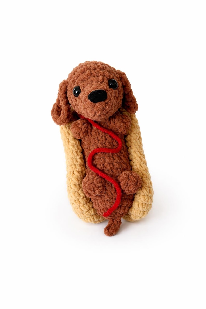 Op de afbeelding: Een handgemaakt bruin gehaakt hondenspeeltje met zwarte ogen en neus, genesteld in een beige gehaakt hotdogbroodje. Een rode gehaakte lijn loopt van de hond. Op een witte achtergrond.