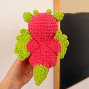 Puede incluir: Un juguete de drag&oacute;n de crochet hecho a mano en colores vibrantes. El drag&oacute;n presenta un cuerpo rosa brillante con alas, cola y detalles de cabeza de color verde lima. El dise&ntilde;o de crochet le da un aspecto suave y texturizado, ideal para ni&ntilde;os.