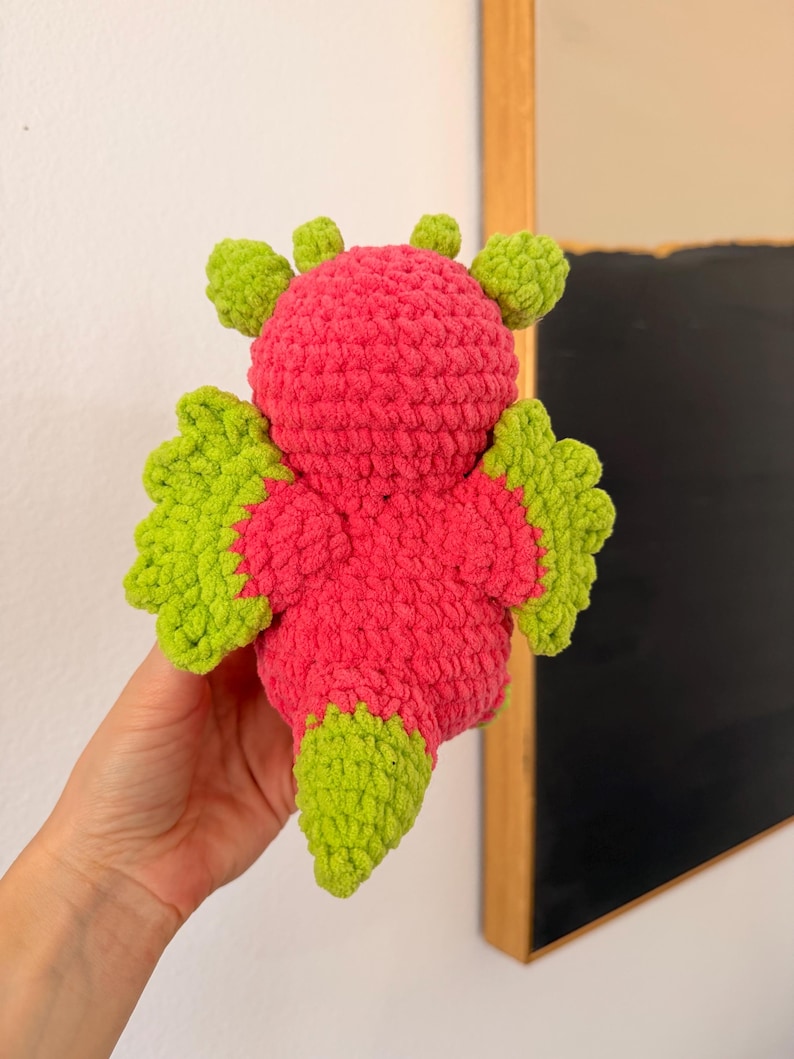 Puede incluir: Un juguete de drag&oacute;n de crochet hecho a mano en colores vibrantes. El drag&oacute;n presenta un cuerpo rosa brillante con alas, cola y detalles de cabeza de color verde lima. El dise&ntilde;o de crochet le da un aspecto suave y texturizado, ideal para ni&ntilde;os.