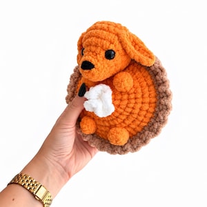May include: A crocheted plush toy shaped like a pumpkin pie slice with a small orange dog head attached to the top. The pie has a bright orange filling, a textured light brown crust edge, and a small white dollop of whipped cream in the center.