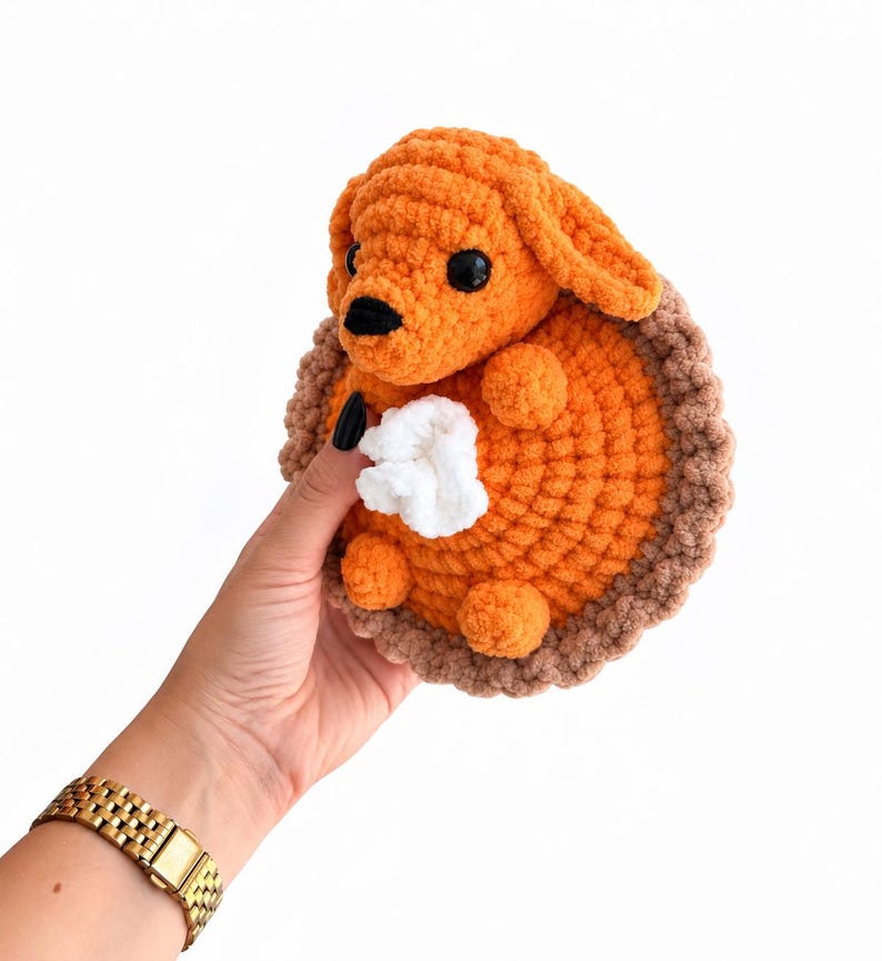 May include: A crocheted plush toy shaped like a pumpkin pie slice with a small orange dog head attached to the top. The pie has a bright orange filling, a textured light brown crust edge, and a small white dollop of whipped cream in the center.