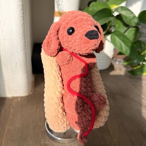 Op de afbeelding: Een handgemaakte gehaakte hond in een hotdogbroodje. De hond is roodbruin van kleur met zwarte ogen en een zwarte neus. Hij heeft een rode lijn. Het hotdogbroodje is beige en lichtbeige. Het speelgoed staat op een zilveren standaard.