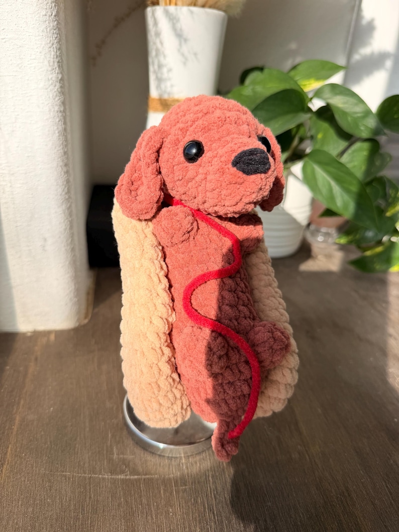 Op de afbeelding: Een handgemaakte gehaakte hond in een hotdogbroodje. De hond is roodbruin van kleur met zwarte ogen en een zwarte neus. Hij heeft een rode lijn. Het hotdogbroodje is beige en lichtbeige. Het speelgoed staat op een zilveren standaard.