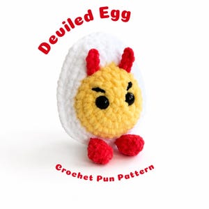 May include: A crocheted plush toy shaped like a hard-boiled egg with a white exterior and a yellow centre. It features two small red horns on top, two red feet at the base, and black plastic eyes. The text Deviled Egg Crochet Pun Pattern is visible.