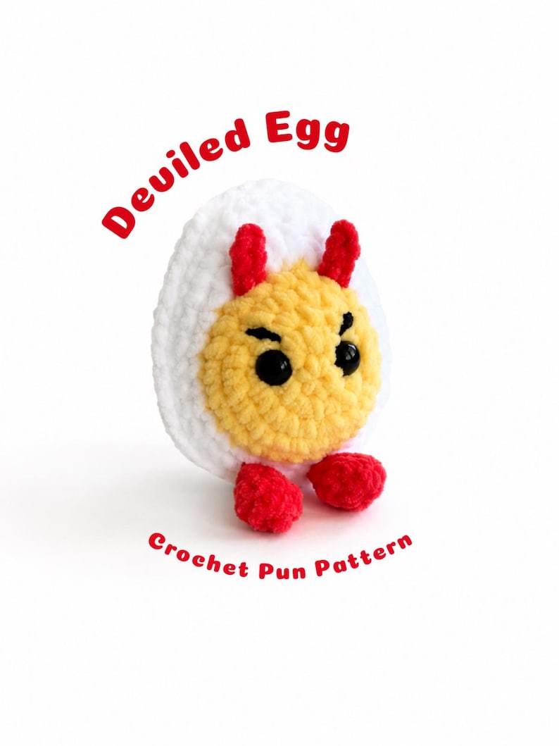 May include: A crocheted plush toy shaped like a hard-boiled egg with a white exterior and a yellow center. It features two small red horns on top, two red feet at the base, and black plastic eyes. The text Deviled Egg Crochet Pun Pattern is visible.