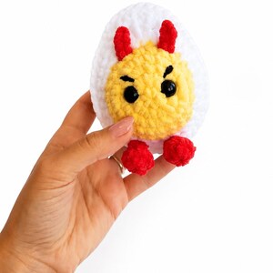 May include: A crocheted plush toy shaped like a hard-boiled egg with a white outer layer and a yellow center. The egg features two small red horns, two red feet, and black plastic eyes with embroidered eyebrows on the yellow portion.
