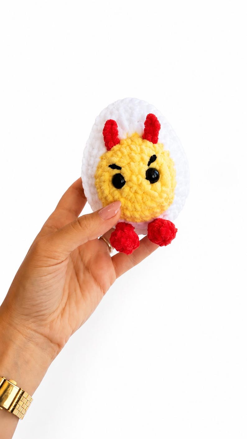 May include: A crocheted plush toy shaped like a hard-boiled egg with a white outer layer and a yellow center. The egg features two small red horns, two red feet, and black plastic eyes with embroidered eyebrows on the yellow portion.