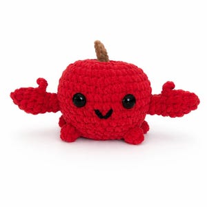 May include: A handmade red crochet apple with a smiling face, black eyes, and outstretched arms. The apple has a brown stem and is made of a soft, textured yarn. A cute and whimsical plush toy.