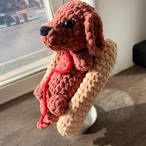 Op de afbeelding: Een handgemaakte gehaakte hond in bruin- en beigetinten, met zwarte ogen en neus. De hond heeft een rood touwtje om de nek en rust op een beige steun. Het pluche speelgoed staat op een houten oppervlak.