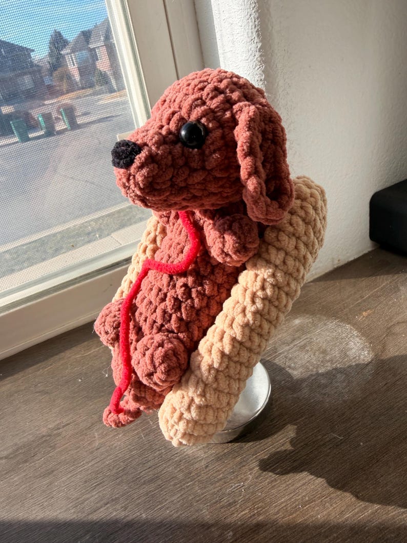 Op de afbeelding: Een handgemaakte gehaakte hond in bruin- en beigetinten, met zwarte ogen en neus. De hond heeft een rood touwtje om de nek en rust op een beige steun. Het pluche speelgoed staat op een houten oppervlak.