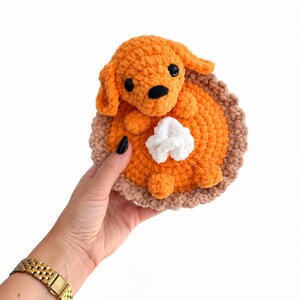May include: A crocheted plush toy shaped like a pumpkin pie with a small orange dog sitting on top. The pie has a light brown crust, a bright orange filling, and a white dollop of whipped cream. The dog has black eyes and a black nose.