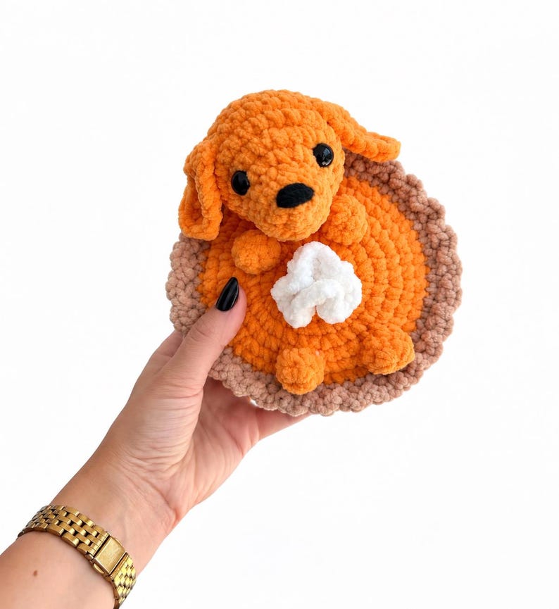 May include: A crocheted plush toy shaped like a pumpkin pie with a small orange dog sitting on top. The pie has a light brown crust, a bright orange filling, and a white dollop of whipped cream. The dog has black eyes and a black nose.